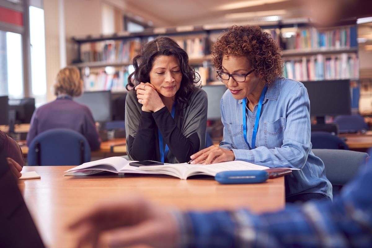 Teacher With Mature Female Adult Student Sitting At Table Working In College Library