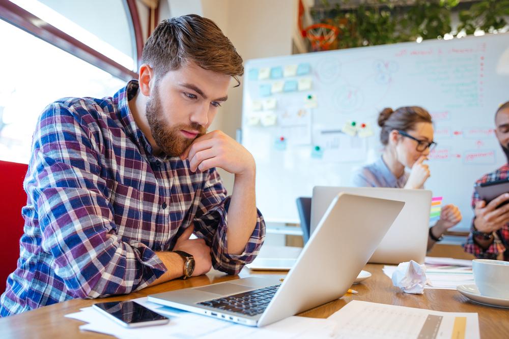 Concentrated bearded young man using laptop while his friends studying together